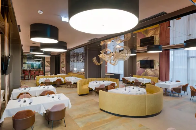 Un restaurant cu mese acoperite cu fețe de masă albe, scaune maronii și banchete galbene dispuse pe un podea cu modele de culori calde.