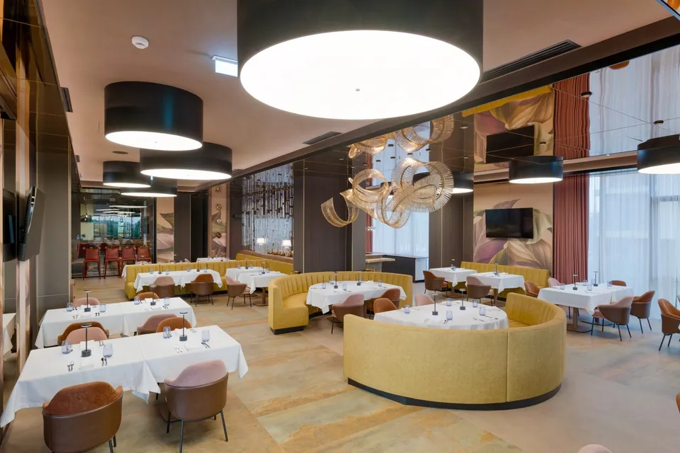 Un restaurant cu mese acoperite cu fețe de masă albe, scaune maronii și banchete galbene dispuse pe un podea cu modele de culori calde.
