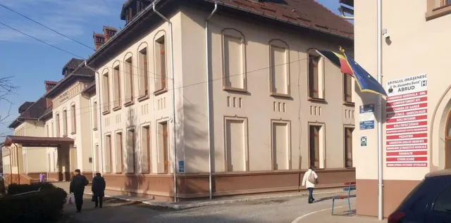 O clădire cu ferestre înalte și două persoane care merg pe un trotuar lângă un perete alb cu un panou roșu.