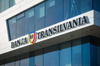 Un semn cu textul Banca Transilvania și un logo colorat este montat pe fațada unei clădiri cu ferestre mari de sticlă.