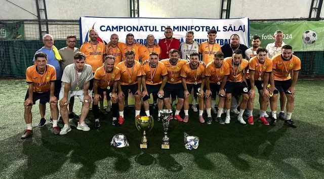 Un grup de bărbați în echipamente sportive portocalii și albe stau în rând pe un teren de fotbal lângă trei trofee și două mingi.