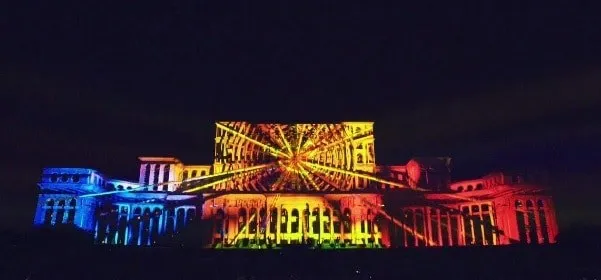 O clădire mare este iluminată de proiecții colorate de tip video mapping în culori galben, portocaliu, roșu și albastru pe un fundal întunecat.