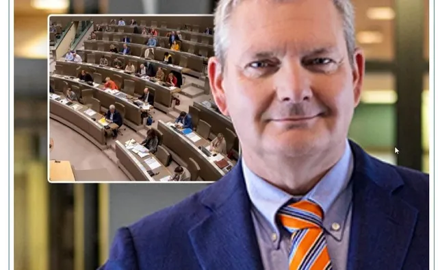 Un bărbat zâmbește, purtând un sacou albastru și o cravată portocalie, cu o imagine a unei săli de ședințe a parlamentului suprapusă în fundal.