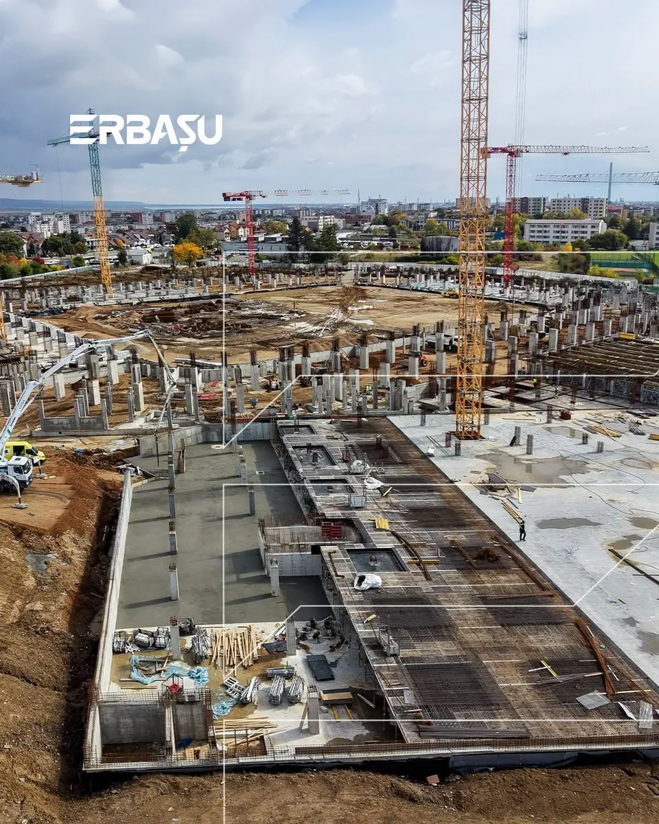 O vedere de sus a unui șantier cu structuri de beton, armătură metalică, macarale și pământ excavat sub un cer înnorat.