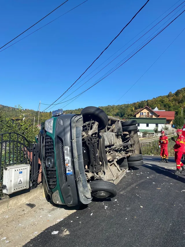 Un autovehicul de culoare verde este răsturnat pe o laterală pe un drum asfaltat, lângă un gard metalic și două persoane în uniforme roșii.