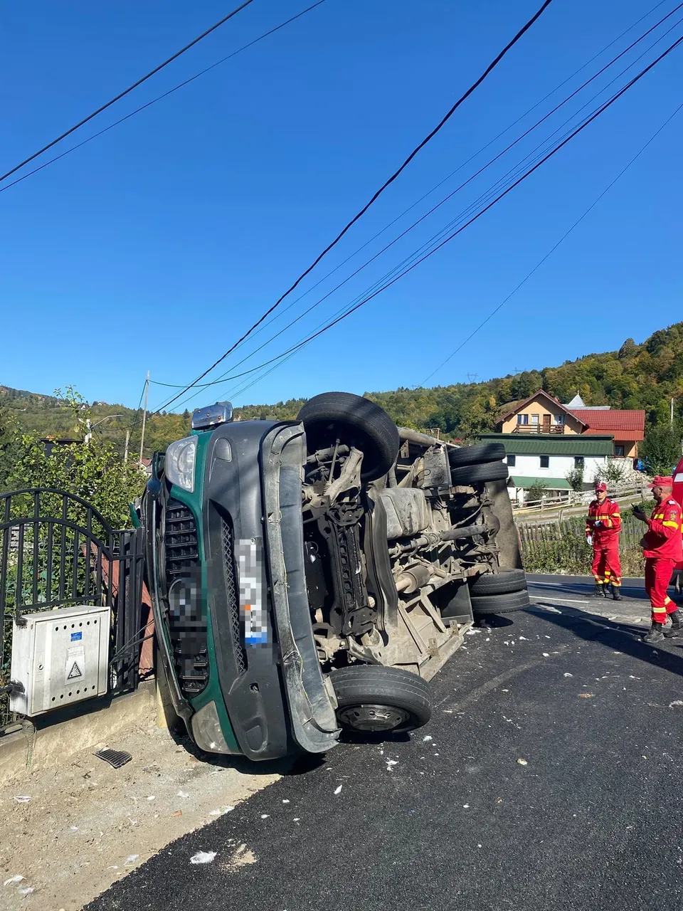 Un autovehicul de culoare verde este răsturnat pe o laterală pe un drum asfaltat, lângă un gard metalic și două persoane în uniforme roșii.