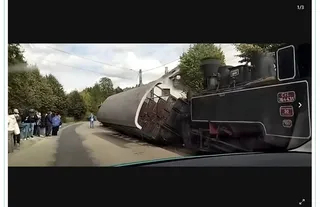 Un vagon de tren răsturnat pe o șină de cale ferată, lângă un drum, cu un grup de oameni stând pe marginea drumului și o locomotivă în fundal.