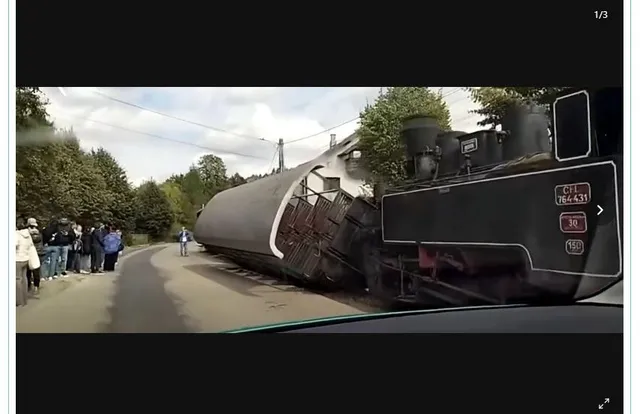 Un vagon de tren răsturnat pe o șină de cale ferată, lângă un drum, cu un grup de oameni stând pe marginea drumului și o locomotivă în fundal.