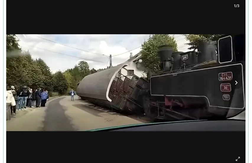 Un vagon de tren răsturnat pe o șină de cale ferată, lângă un drum, cu un grup de oameni stând pe marginea drumului și o locomotivă în fundal.