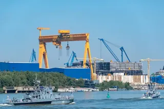 Două nave mici navighează pe un râu în fața unui șantier naval cu macarale galbene și albastre și structuri industriale sub un cer senin.