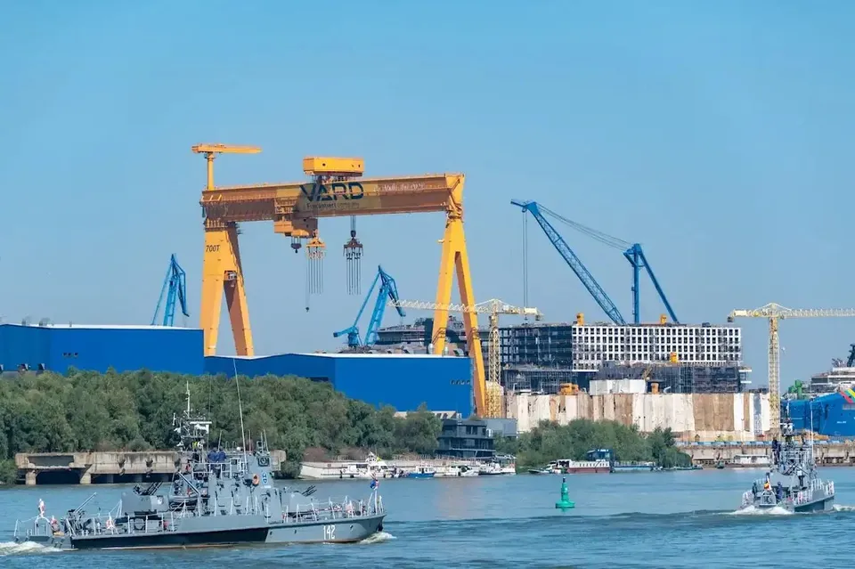Două nave mici navighează pe un râu în fața unui șantier naval cu macarale galbene și albastre și structuri industriale sub un cer senin.