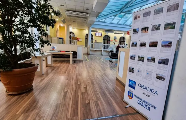 Un panou informativ cu fotografii și text stă pe un podea de lemn într-un spațiu interior cu un tavan de sticlă și o plantă în ghiveci.