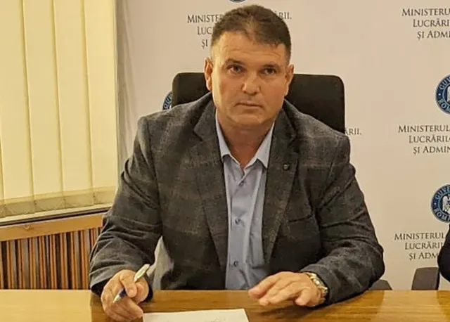 Un bărbat cu sacou gri și cămașă deschisă stă la o masă de lemn în fața unui fundal alb cu text.