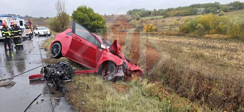 Un autoturism roșu este răsturnat pe o marginea drumului, lângă un motor separat, în timp ce personalul de intervenție se află lângă o ambulanță.