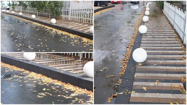 Un trotuar îngust cu bordură înaltă și pietre rotunde albe este situat lângă un gard alb și o stradă umedă cu frunze căzute.