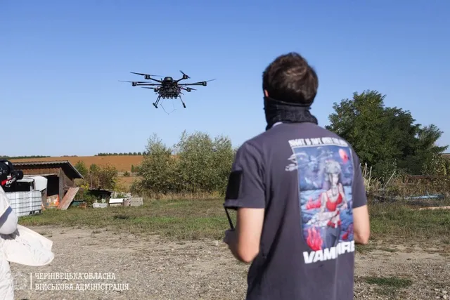 Un bărbat cu un tricou gri stă cu spatele, privind un quadcopter care zboară pe cerul senin deasupra unui teren cu vegetație și un copac.