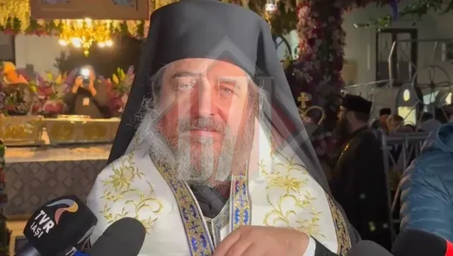 Un bărbat cu barbă și pălărie neagră poartă haine albe cu broderii aurii și este încadrat de microfoane în prim-plan.