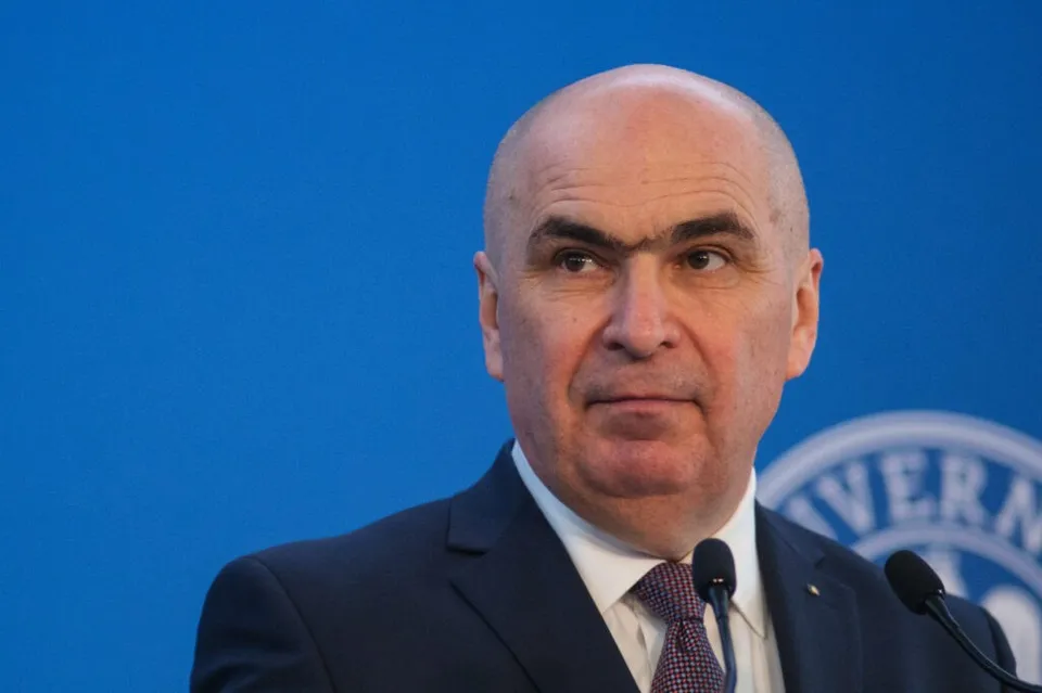 Un bărbat cu părul rare și sprâncene groase poartă un costum închis, cămașă albă și cravată, stând în fața unui fundal albastru cu microfoane.