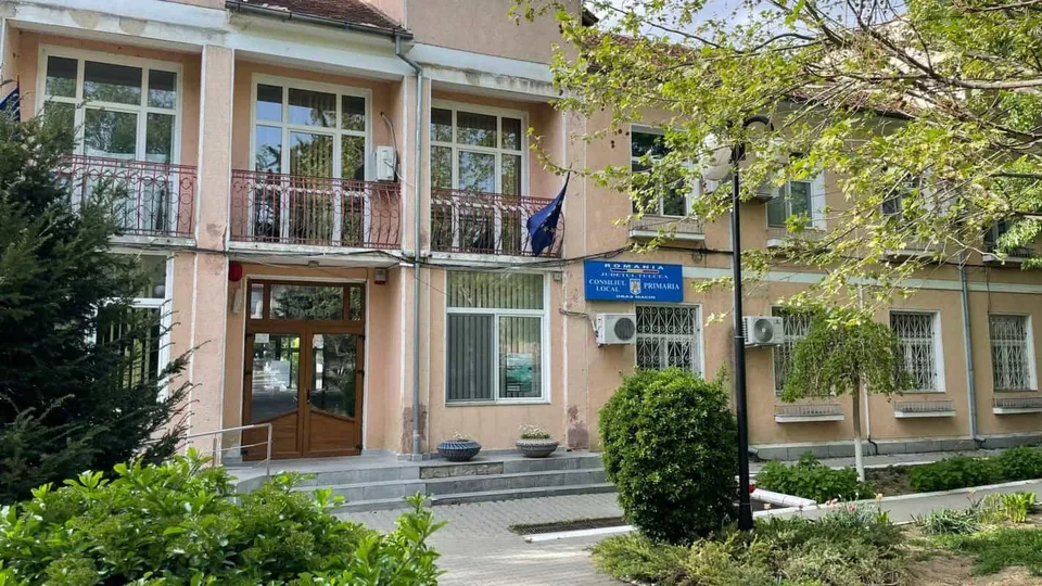 O clădire cu două etaje, de culore deschisă, cu ferestre albe, balustrade metalice la etaj, o ușă de intrare din lemn și o plăcuță albastră pe perete.