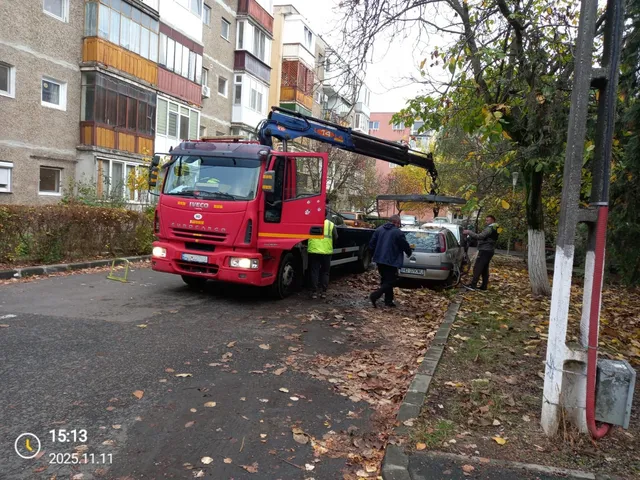 Un camion roșu cu braț hidraulic este parcat pe un drum, lângă un autoturism gri și câțiva oameni în apropiere.