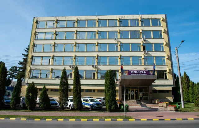 O clădire de patru etaje cu ferestre rectangulare este situată în spatele unor copaci înalți și verzi, lângă un parcare cu mașini albe și albastre.