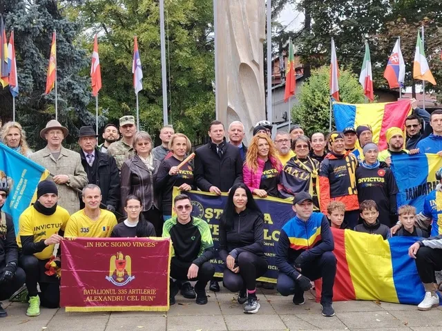 Un grup de oameni stă strâns pentru o fotografie de grup, purtând haine colorate și ținând diverse steaguri și un banner dreptunghiular.