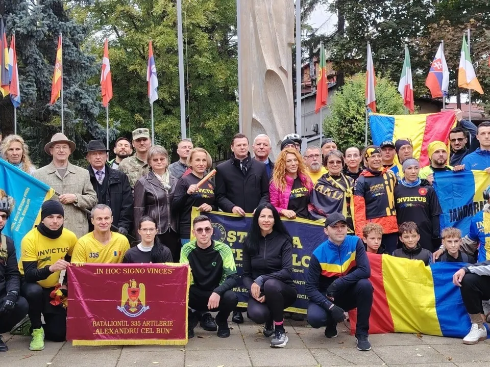 Un grup de oameni stă strâns pentru o fotografie de grup, purtând haine colorate și ținând diverse steaguri și un banner dreptunghiular.