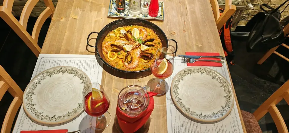 O tavă neagră cu paella, garnitură de lămâie și fructe de mare, este așezată pe o masă de lemn lângă două farfurii și pahare cu băuturi.