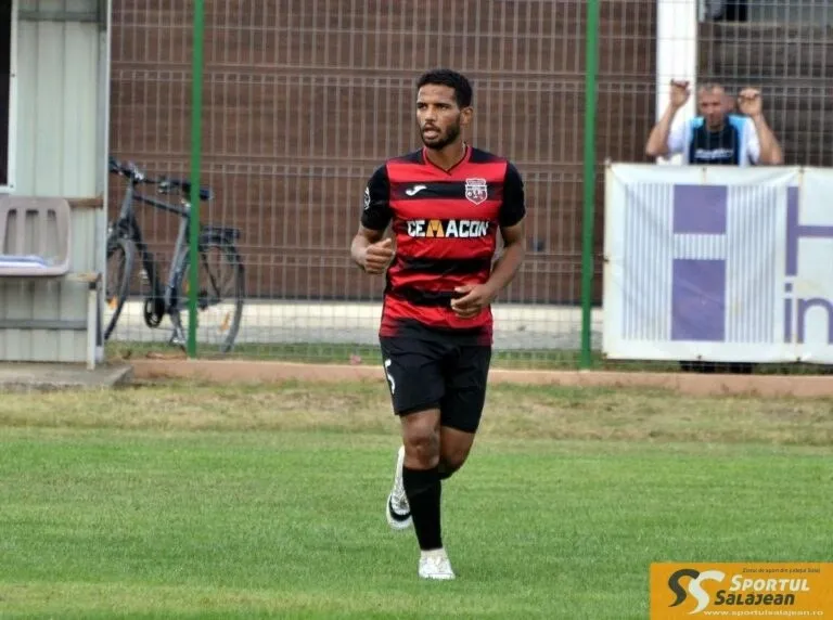 Un bărbat cu barbă poartă un tricou roșu și negru cu textul CEMACON pe un teren de fotbal acoperit cu iarbă.