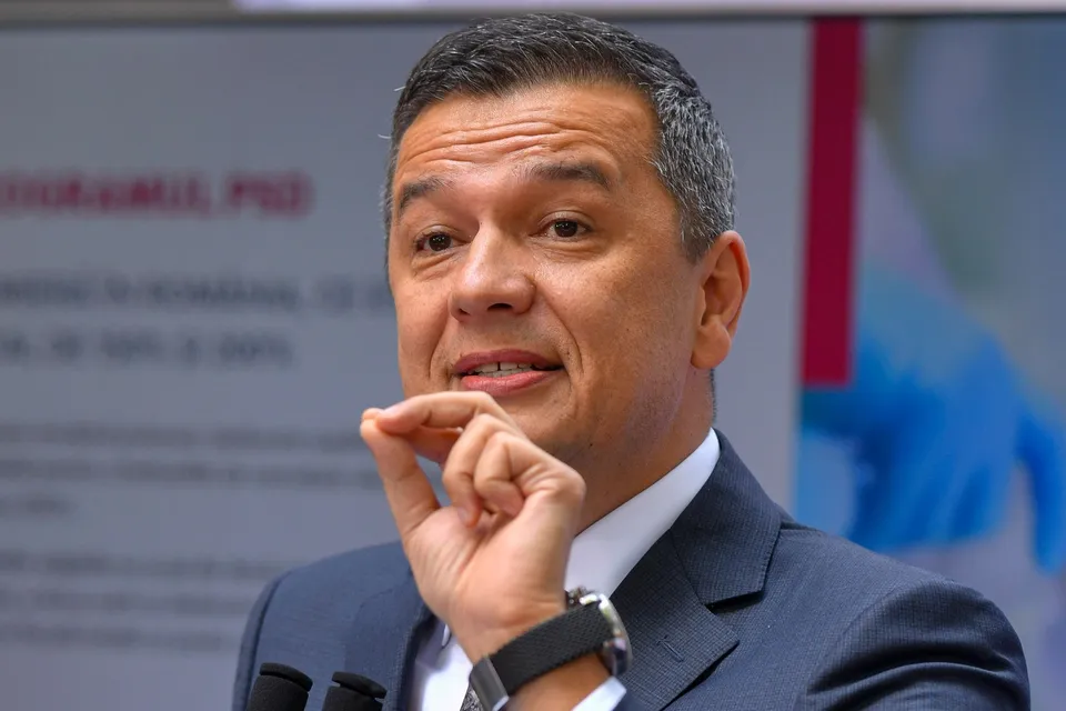 Un bărbat cu părul scurt și cămașă albă poartă un sacou închis la culoare și ridică mâna dreaptă lângă față.