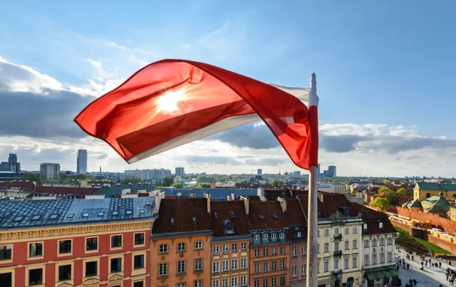 O steag polonez roșu și albagiu flutură pe un stâlp deasupra unor clădiri colorate sub un cer cu nori.