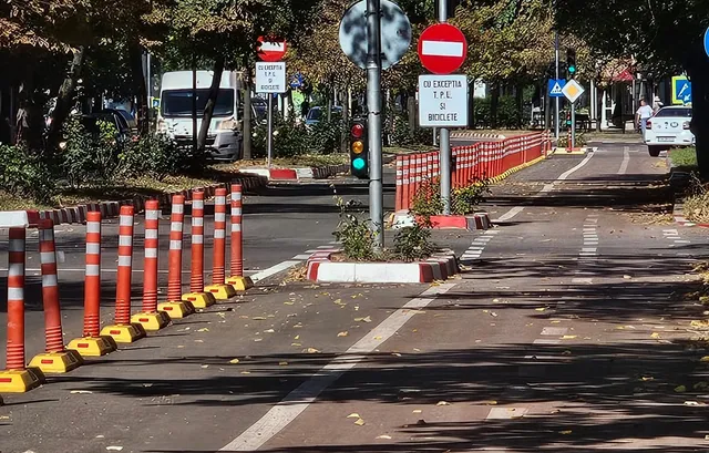O pistă de biciclete asfaltată este delimitată de conuri de trafic portocalii și albe, având semne de circulație și un vehicul alb în fundal.