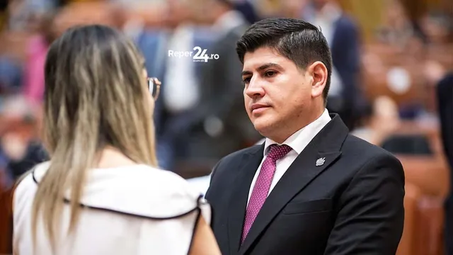 Un bărbat cu părul scurt și negru, purtând un costum întunecat și o cravată roz, privește spre o femeie cu părul blond văzută din spate.