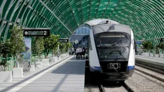 Un tren alb și albastru stă pe un binar lângă un peron cu copaci în plante și un acoperiș de sticlă verde.