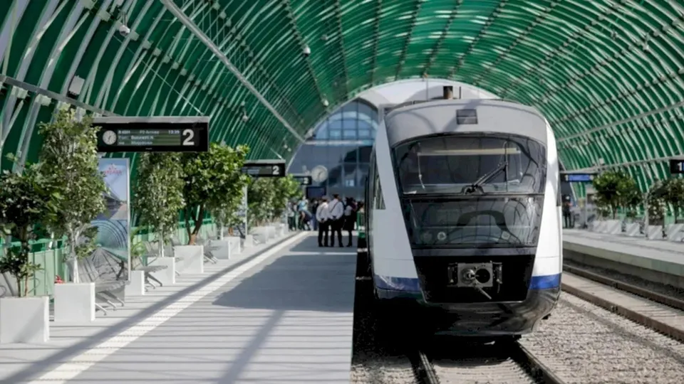Un tren alb și albastru stă pe un binar lângă un peron cu copaci în plante și un acoperiș de sticlă verde.