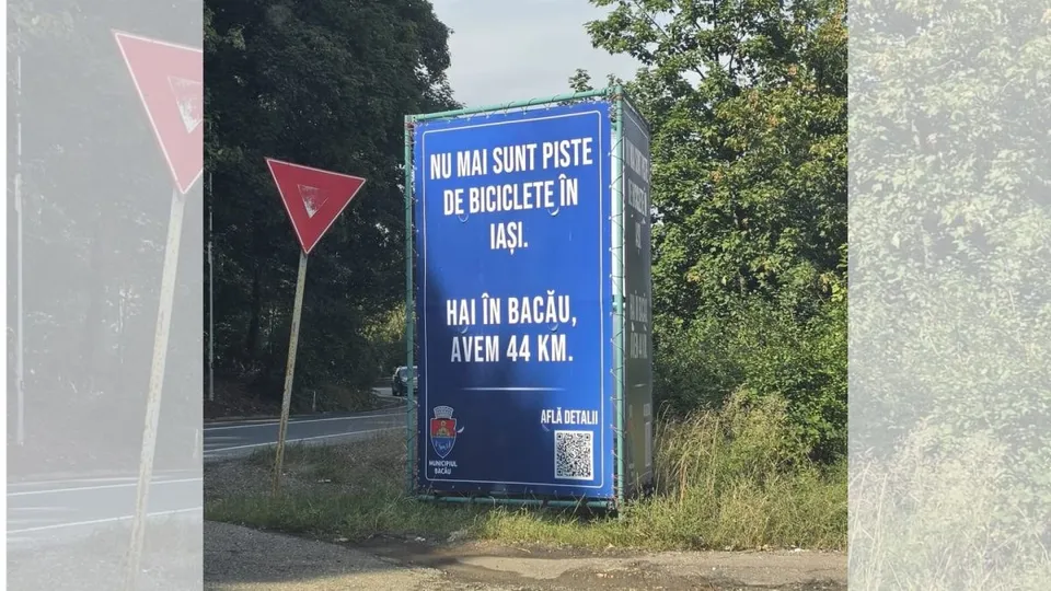 Un panou publicitar albastru cu text alb și un cod QR este amplasat pe marginea unui drum lângă vegetație.