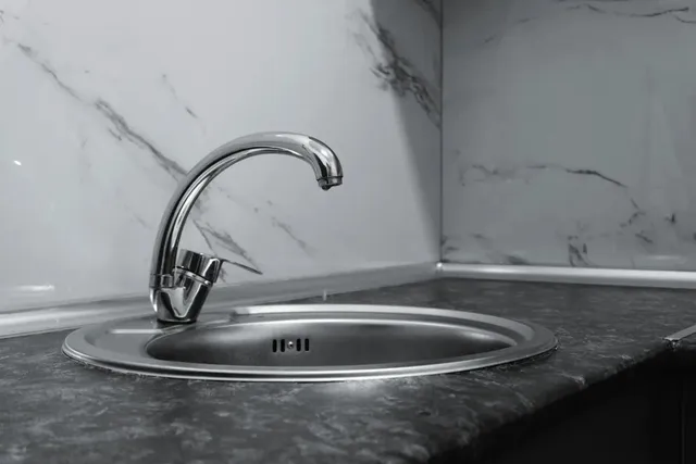 Un robinet metalic este montat pe o chiuvetă de inox într-o bucătărie cu blat și perete de marmură.