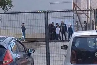 Un grup de persoane stă adunat în spatele unui gard metalic, lângă două mașini parcate în prim-plan.