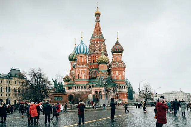 O catedrală cu cupole colorate se ridică deasupra unei piețe aglomerate cu oameni care merg sau stau în picioare sub un cer înnorat.