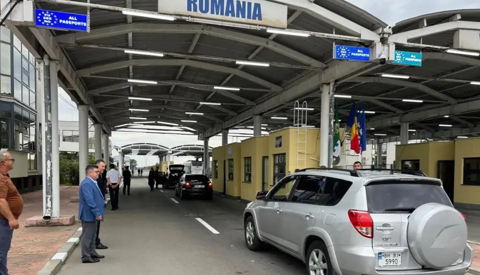 Mașini circulă pe o șosea de asfalt sub o structură metalică de acoperire, lângă clădiri galbene și câteva persoane stând pe marginea drumului.