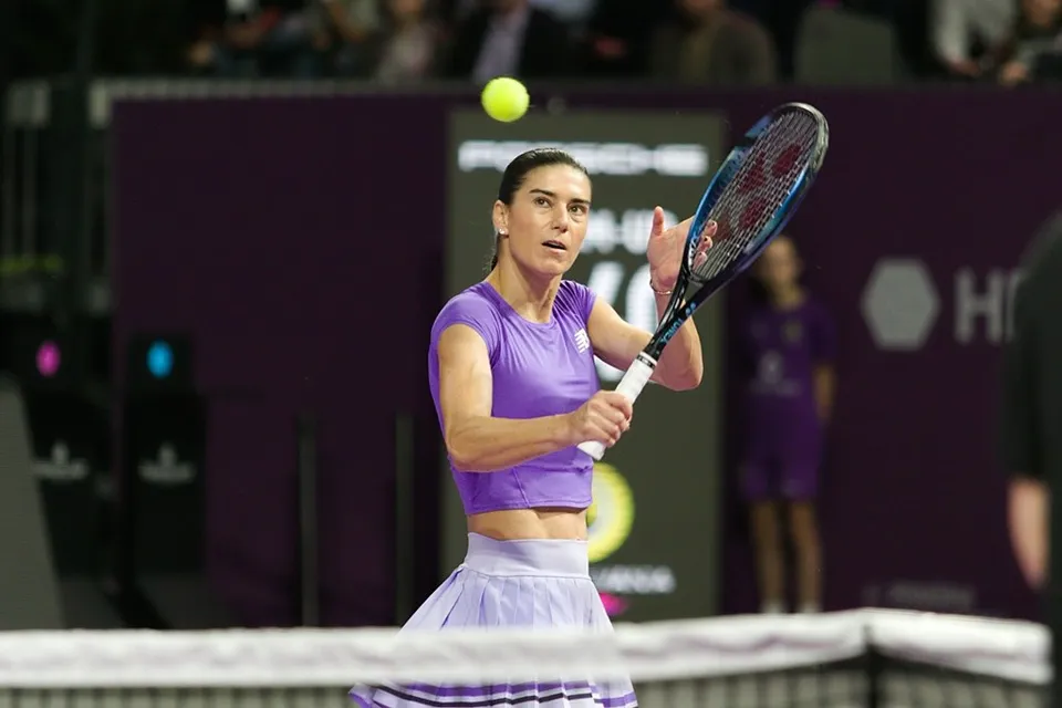 O femeie cu o cămașă violetă și o fustă albă lovește o minge de tenis cu o rachetă pe un teren de tenis.