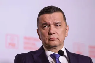 Un bărbat cu părul scurt, purtând sacou închis la culoare, cămașă albă și cravată albastră, stă în fața unui microfon pe un fundal deschis la culoare.