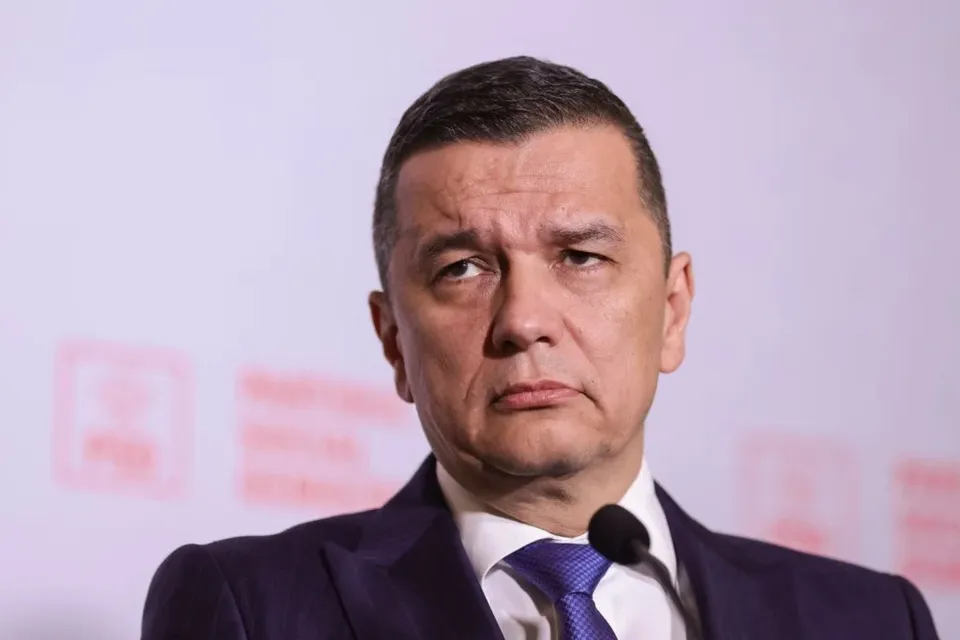 Un bărbat cu părul scurt, purtând sacou închis la culoare, cămașă albă și cravată albastră, stă în fața unui microfon pe un fundal deschis la culoare.