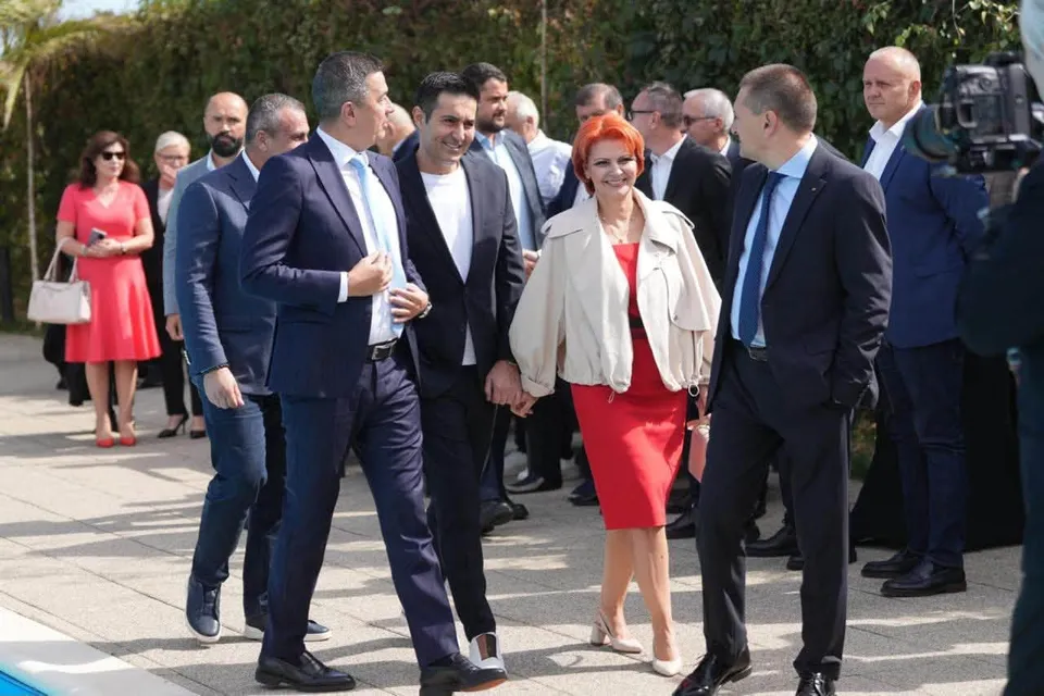 Un grup de persoane îmbrăcate în costume și haine casual merge pe o cărare pavată lângă vegetație sub lumina soarelui.