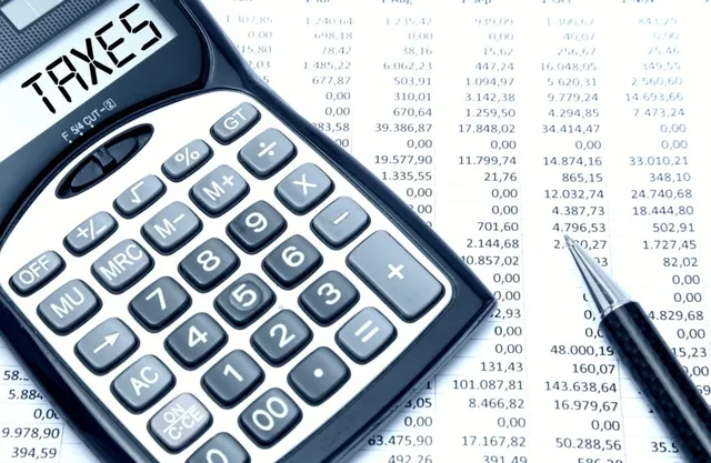 O poză de aproape a unei calculatoare electronicice afișând cuvântul TAXES, alăturate de un pix și a unui tabel cu date financiare.