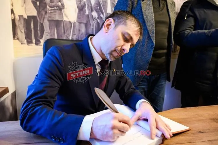 Un bărbat cu sacou și cravată stă la o masă de lemn și scrie într-un caiet deschis cu un pix.