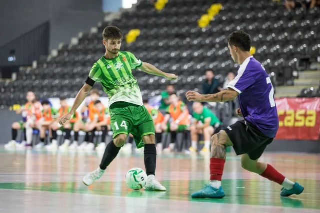 Un jucător în echipament verde și alb controlează un balon pe un teren de futsal în fața unui adversar îmbrăcat în violet și negru.