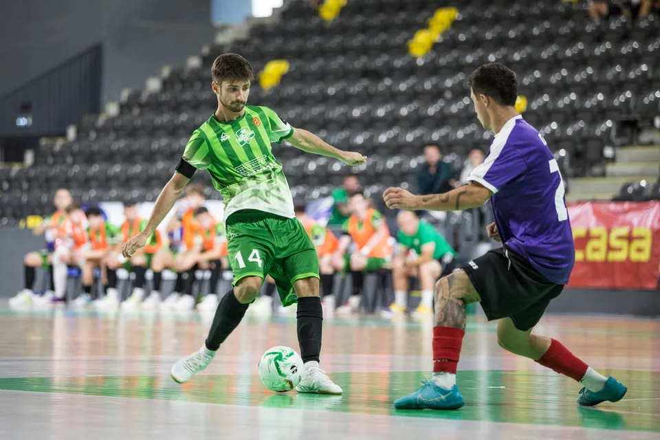 Un jucător în echipament verde și alb controlează un balon pe un teren de futsal în fața unui adversar îmbrăcat în violet și negru.