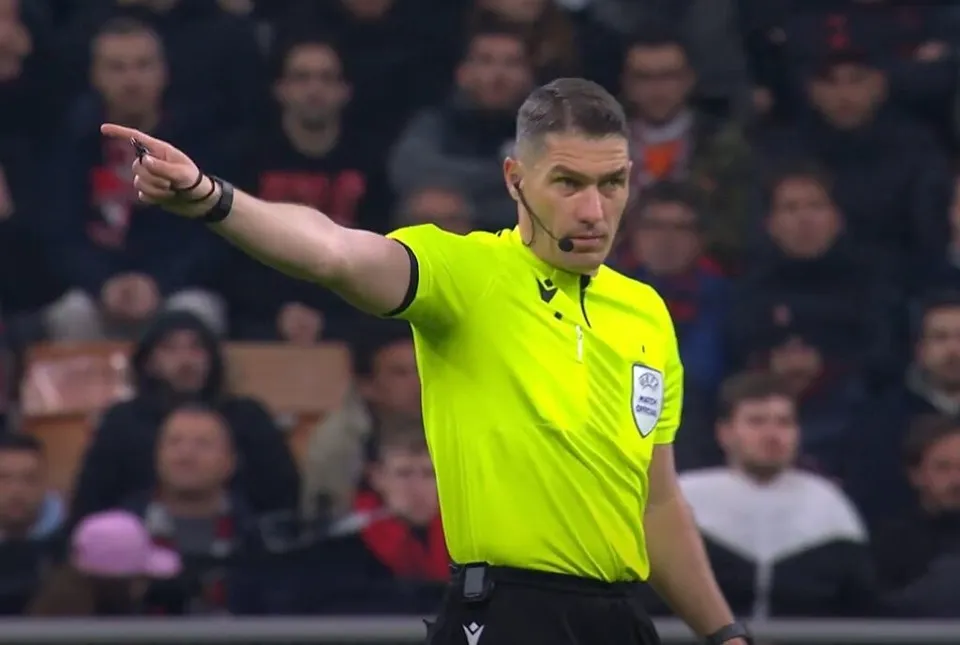 Un bărbat cu tricou galben și căști de comunicare arată spre stânga cu degetul arătător întins într-un stadion cu spectatori în fundal.