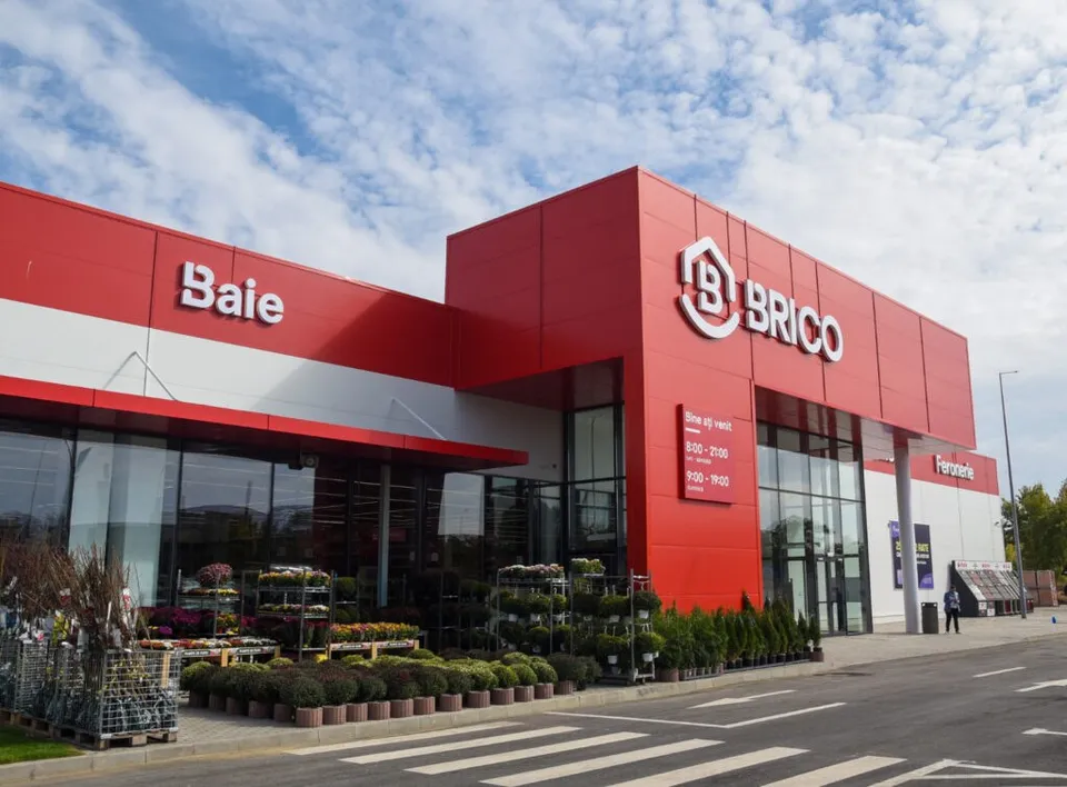 O clădire comercială modernă cu fațadă roșie prezintă semnele BRICO și Baie, având plante și rafturi cu produse expuse în fața vitrinelor mari.